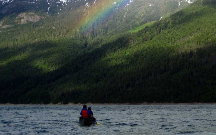 a canoe sits on a body of water facing a green mountain face with a rainbow cast in front of it